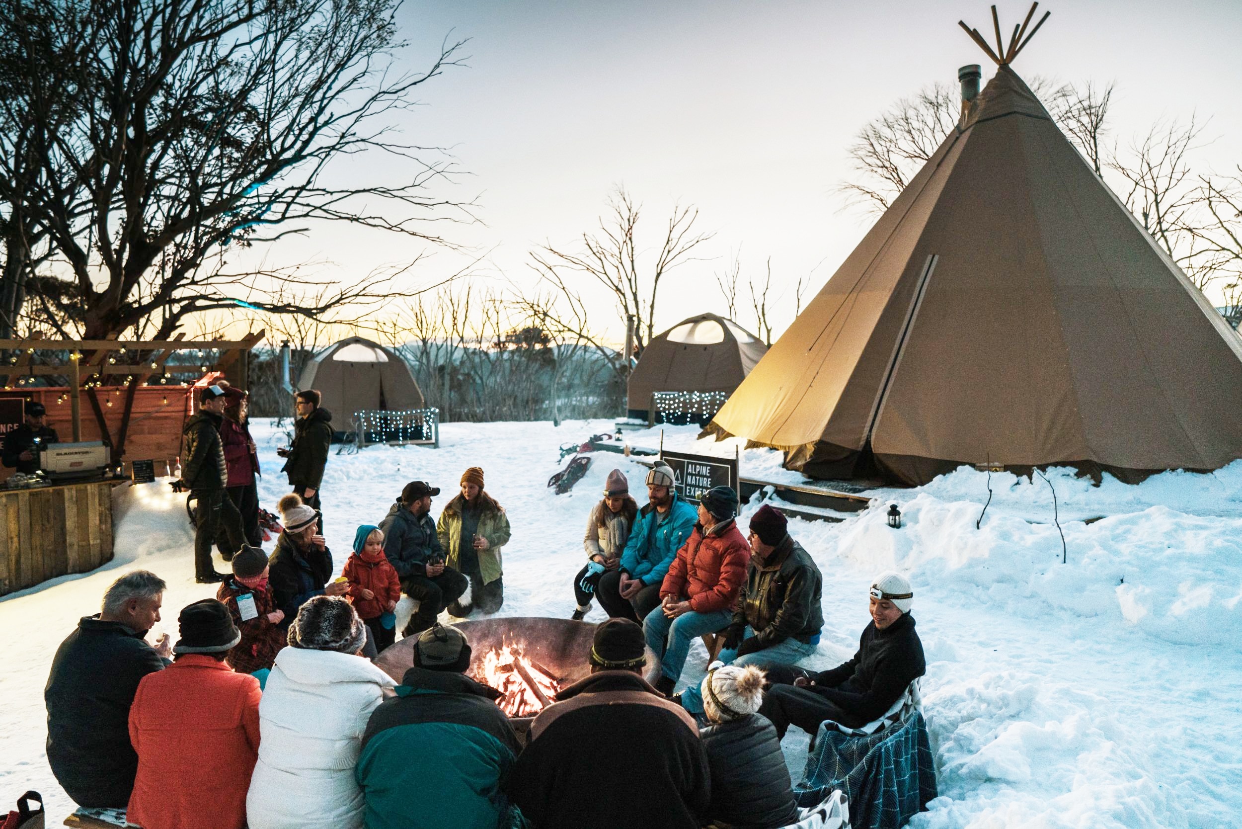 People sit around a fire in the snow with a tent in the background. 