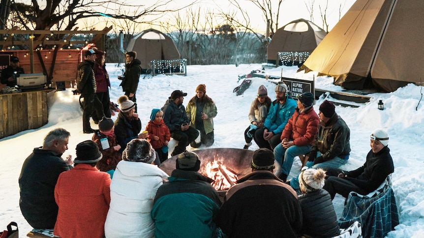 People sit around a fire in the snow with a tent in the background. 