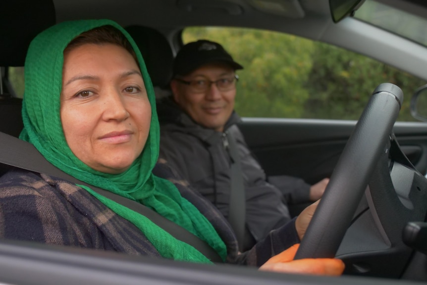 A woman in a green headscarf sitting behind the wheel in a car, while a man in the passenger seat smiles to camera too