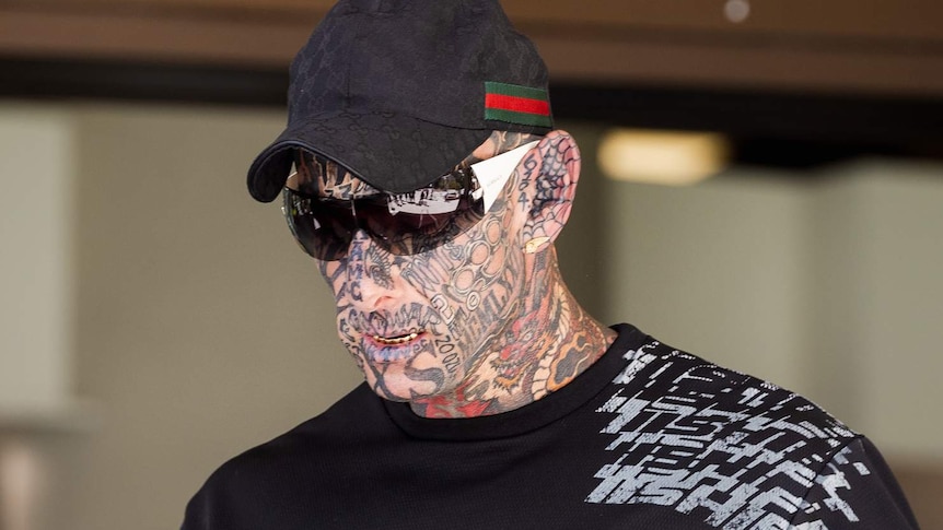 Brajkovich walks wearing a hat and sunglasses. He has full face tattoos and appears to have gold caps on most of his teeth.