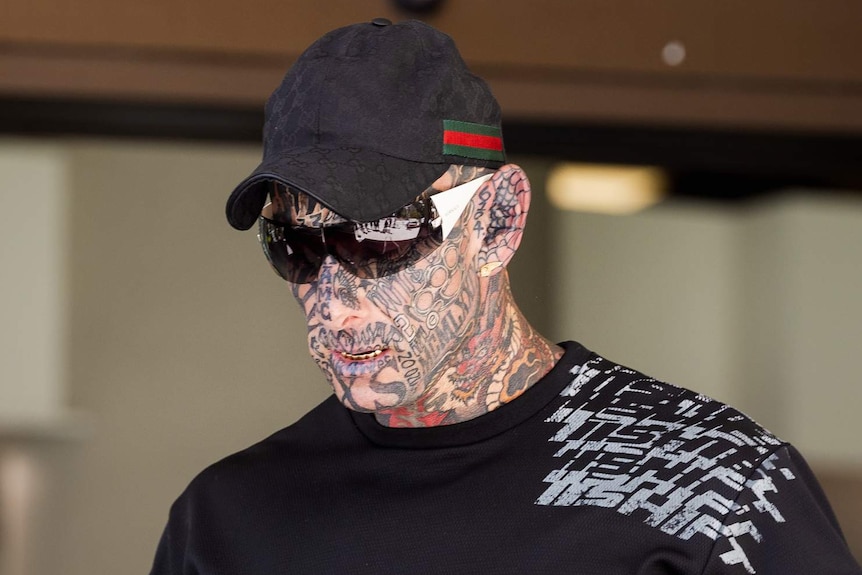 Brajkovich walks wearing a hat and sunglasses. He has full face tattoos and appears to have gold caps on most of his teeth.