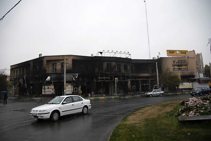 A white car drives away from a two-storey building that is blackened with ash and smoke