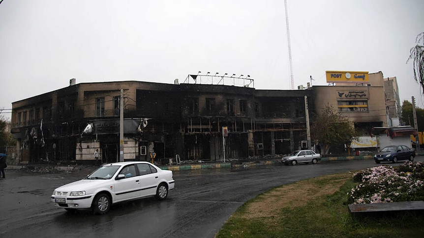 A white car drives away from a two-storey building that is blackened with ash and smoke