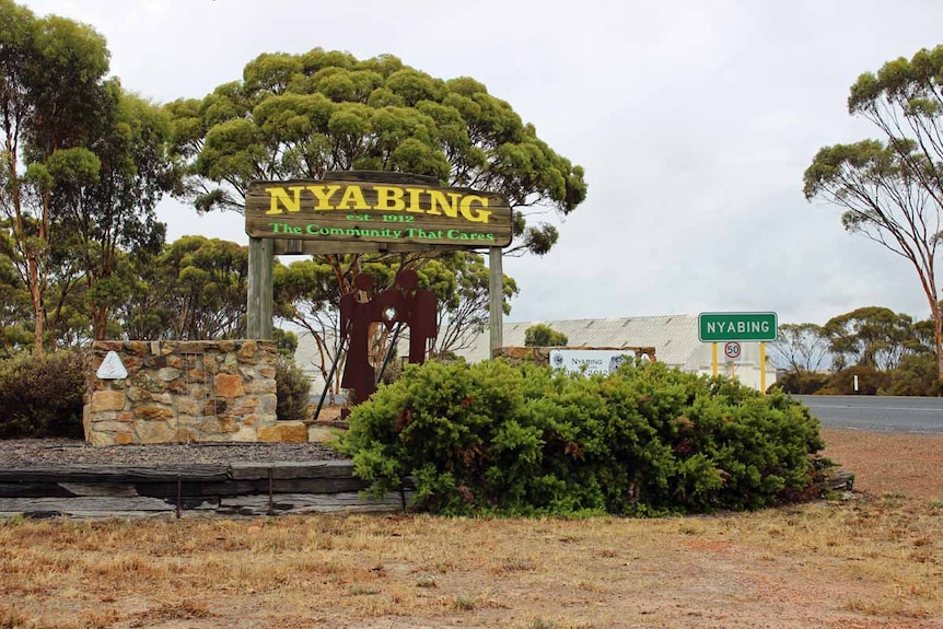 A town sign situated next to a road and near huge grain storage bins.