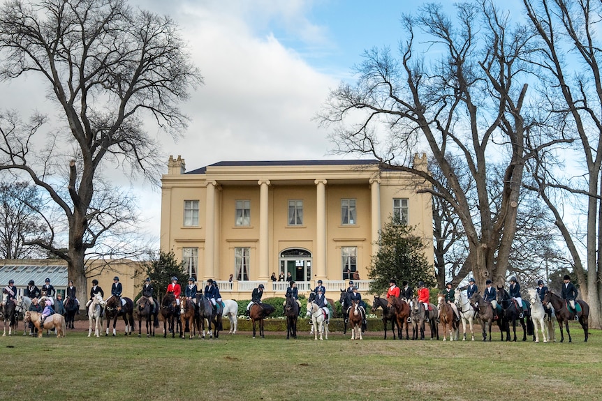 Dozens of equestrians wearing navy, red and black blazers line up outside a daffodil-coloured mansion on a partly cloudy day.