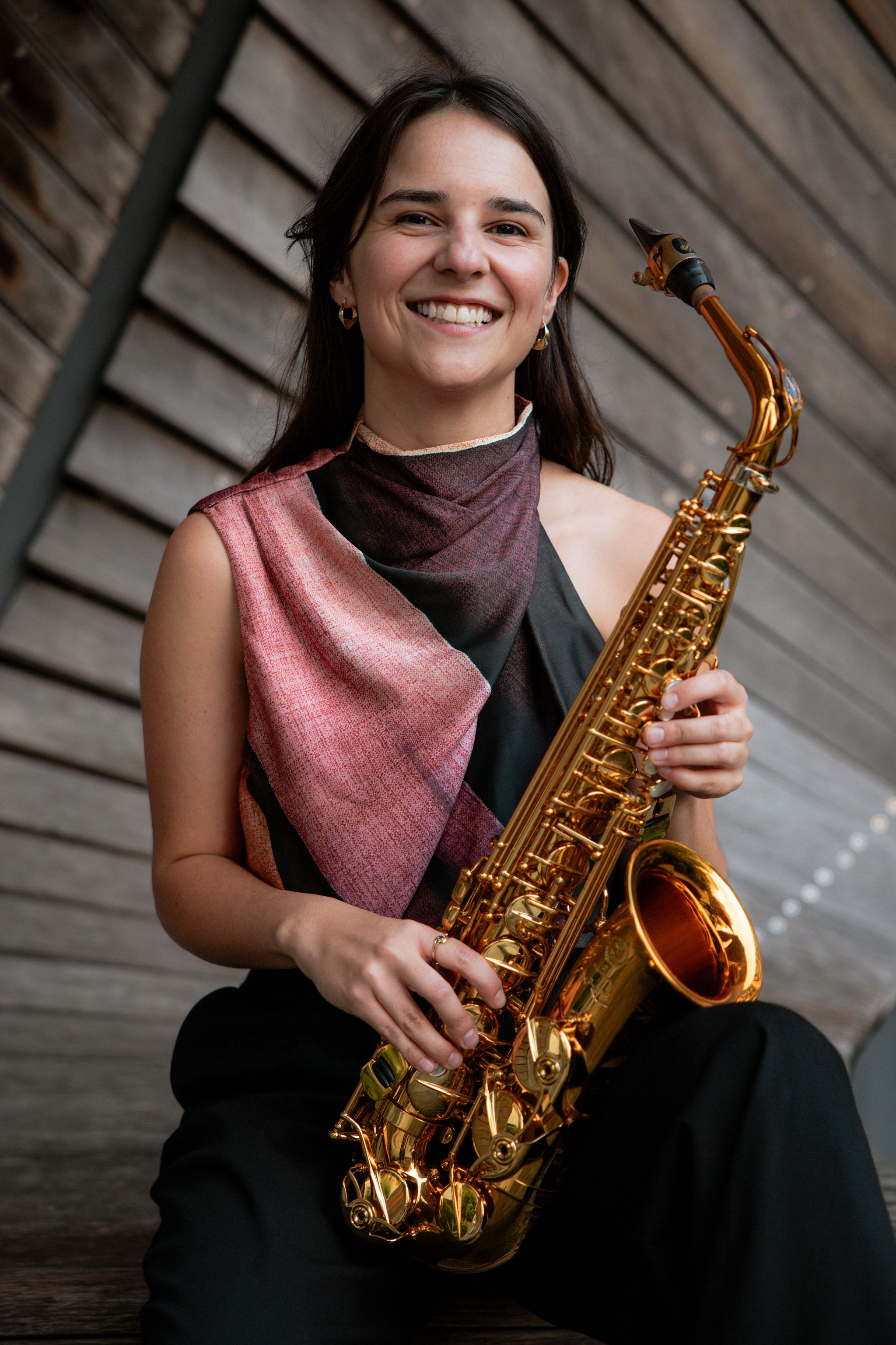 Portrait of a smiling Alice Morgan seated with a gold alto saxophone against angled wooden panels.