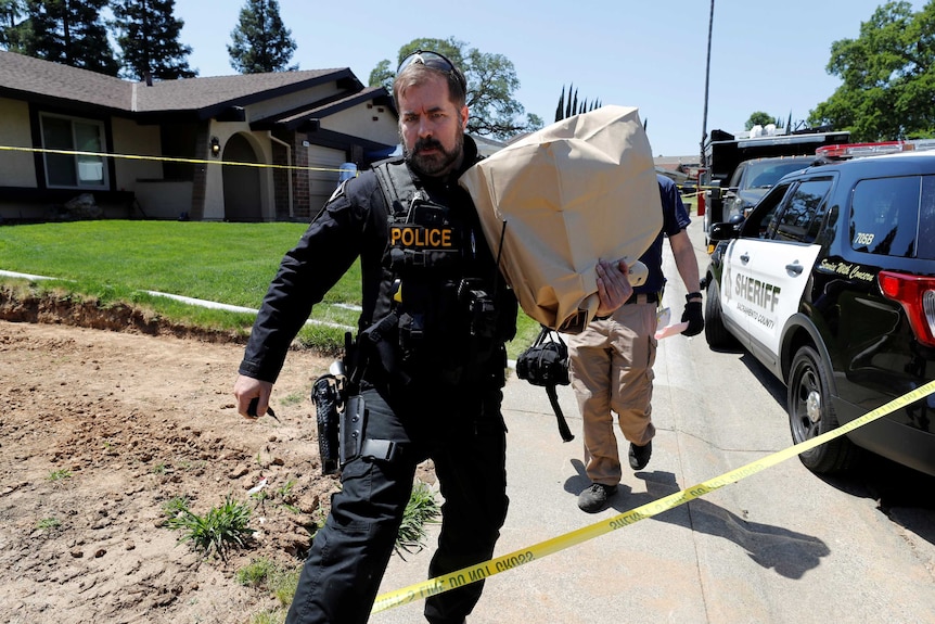 A police officer holds a brown paper bag outside a house