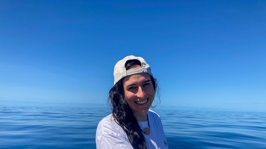 A woman in a white shirt and backwards cap smiles in front of pure, blue ocean.