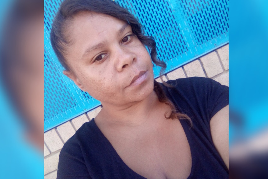 A dark-skinned woman in a black tee-shirt takes a selfie in front of a blue fence.