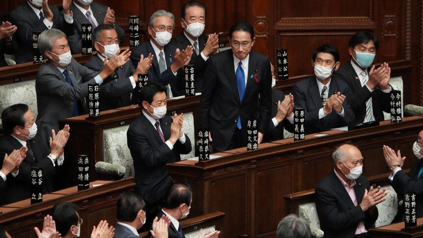 An Asian man in dark suit and blue tie stands silently as face masked men in suits applaud him in political chamber.