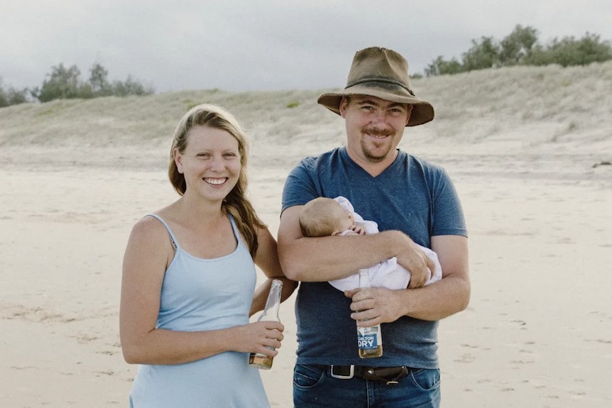 a woman and a man holding a baby stand next to each other smiling on a beach, they are each holding a beer bottle