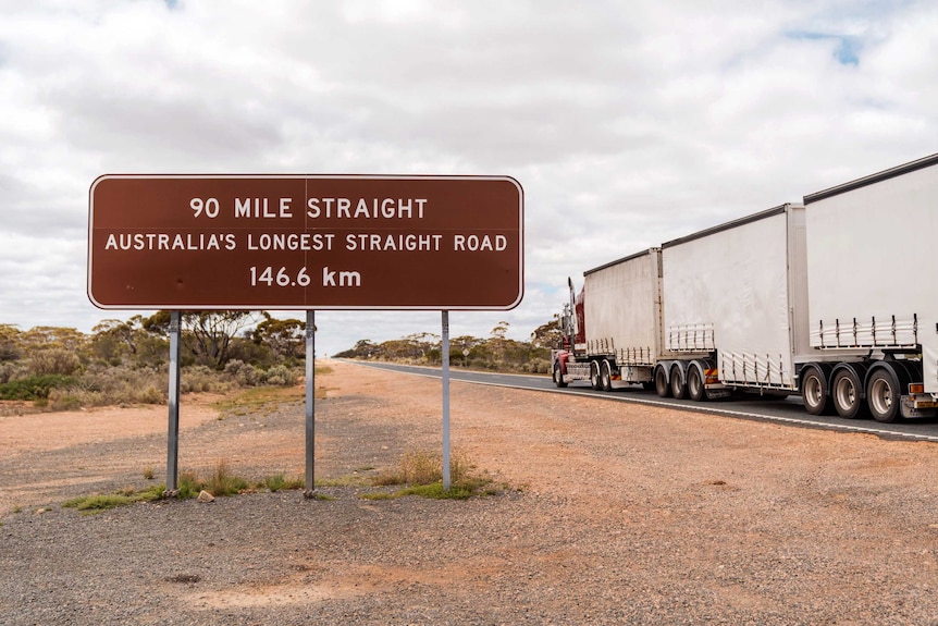 A road train passing a sign proclaiming '90 Mile Straight'.