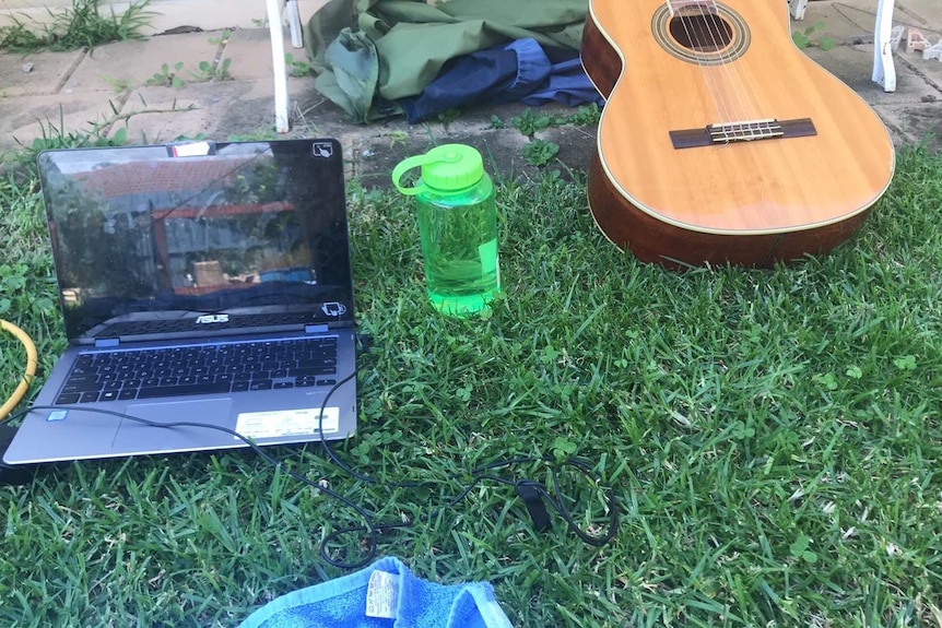 A laptop is seen on the grass, with a bottle of water and guitar.