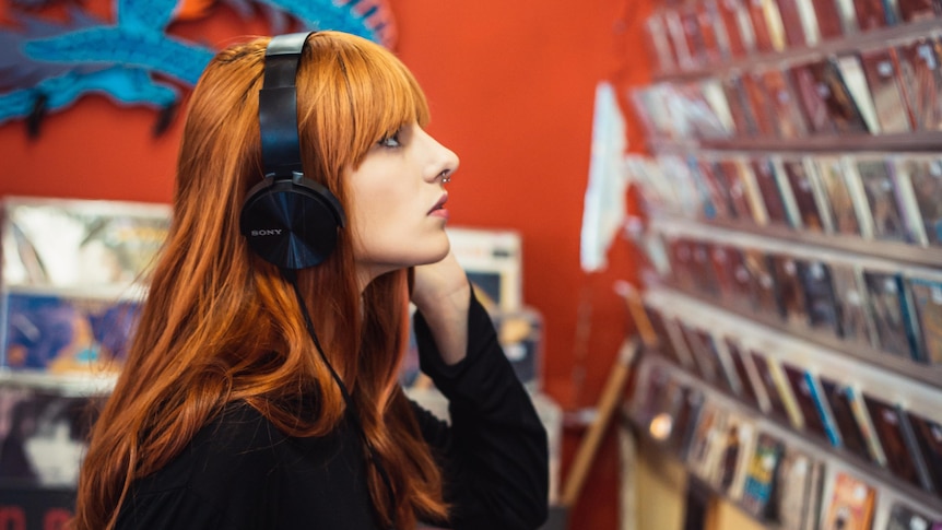 A woman with red hair wearing headphones stares at a wall of CDs in a shop