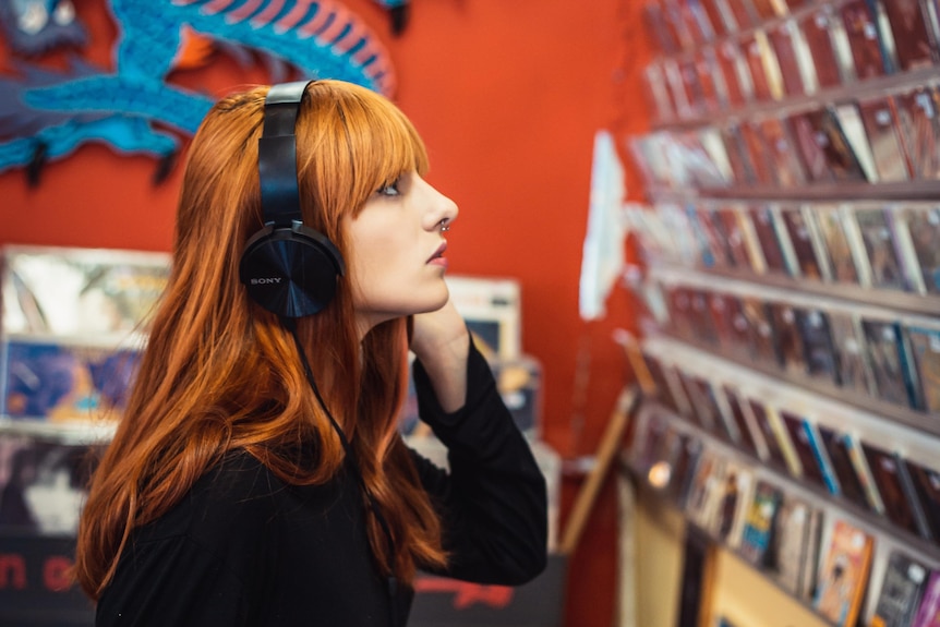 A woman with red hair wearing headphones stares at a wall of CDs in a shop