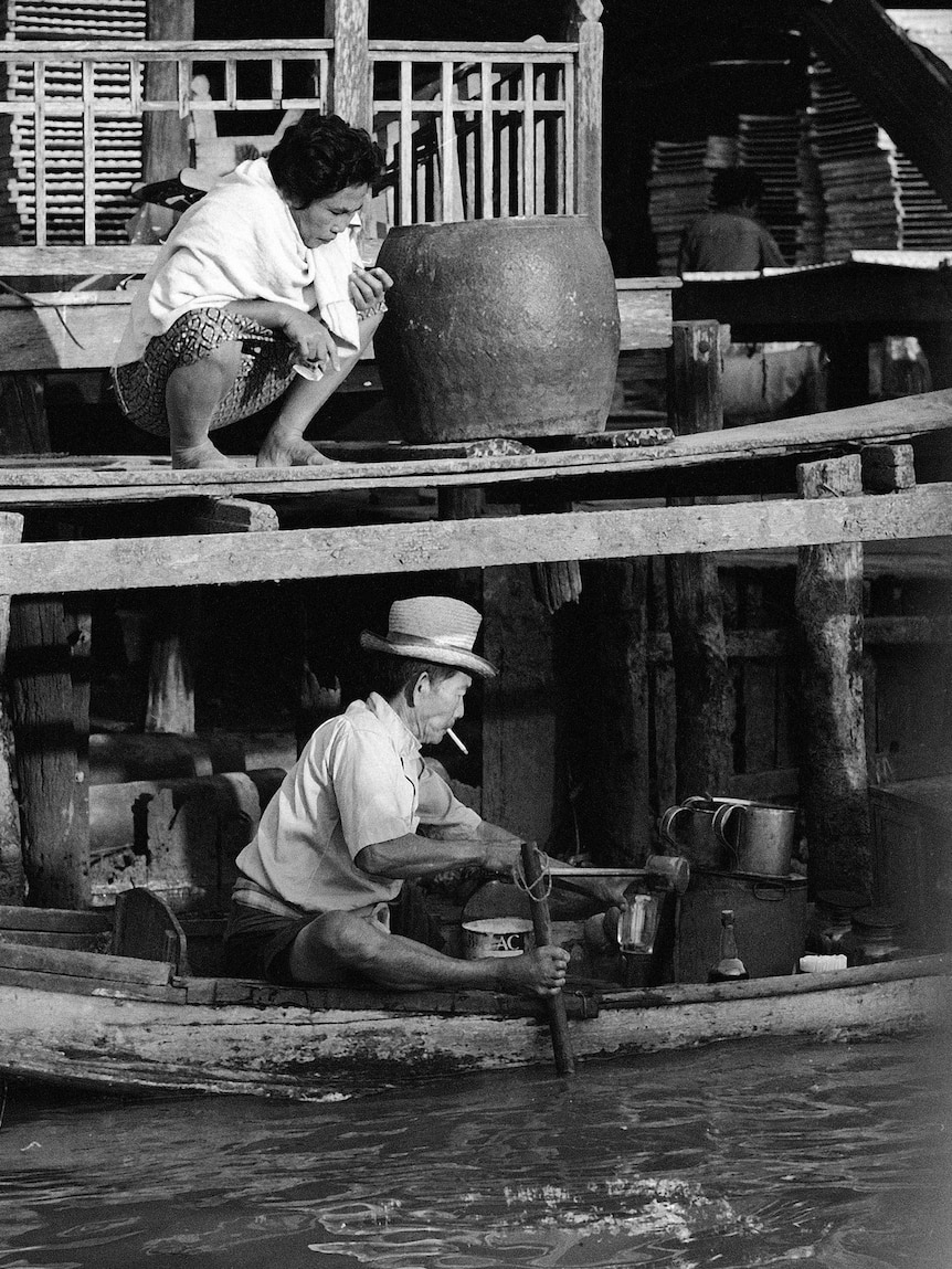 A woman crouches on a wooden platform above a canal as a boat passes with a man smoking a cigarette and steering with his foot