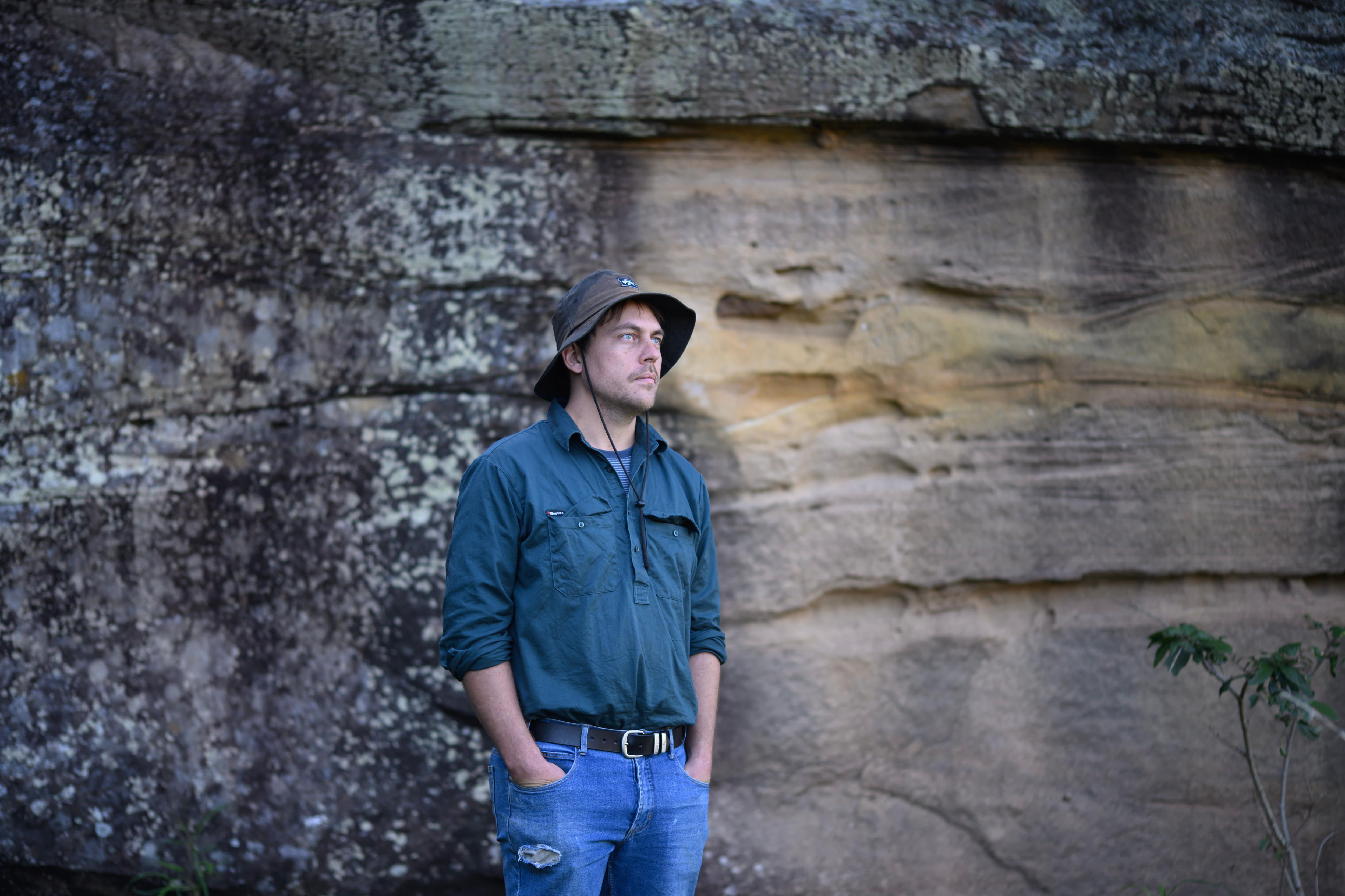 A man stands with his hands in his pockets in front of a sandstone cliff face.