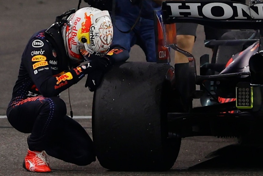 a formula 1 racer kneels down with his head on the tyre of his car. he looks emotional