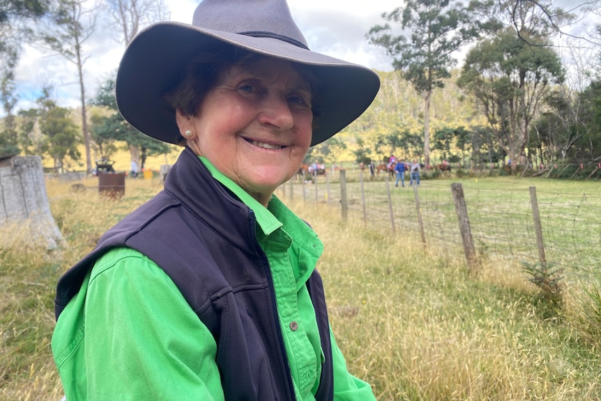 A woman in a green shirt, navy vest and Broad-brimmed hat sits on a log. 