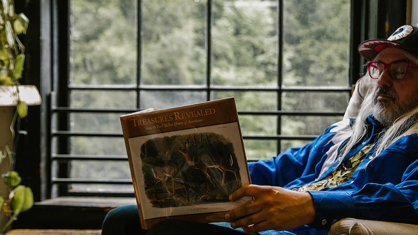 J Mascis sitting with his eyes closed, holding a picture book entitled Treasures Revealed