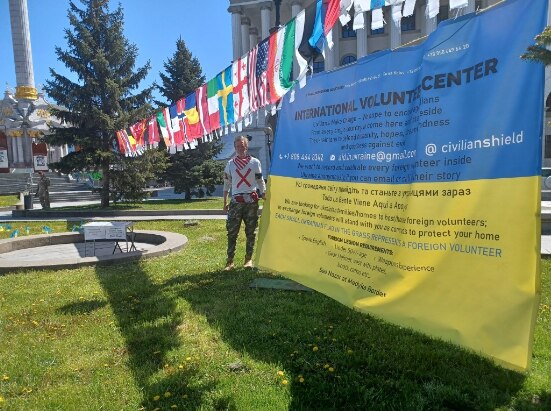 A man wearing a white t-shirt with a red cross on it standing next to a large ukrainian flag covered in text