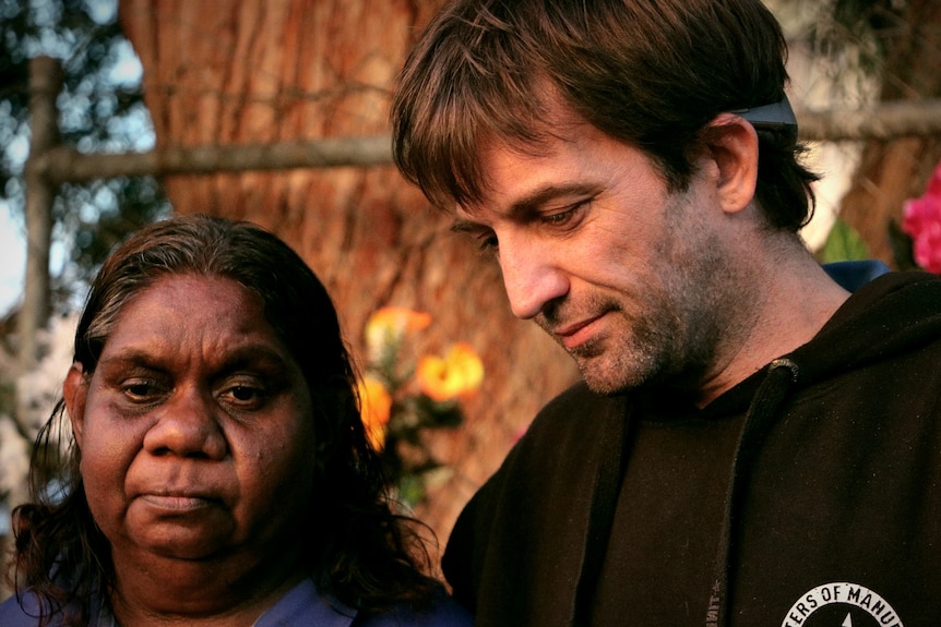 A white man looking down with his arm around an Aboriginal woman looking solemn