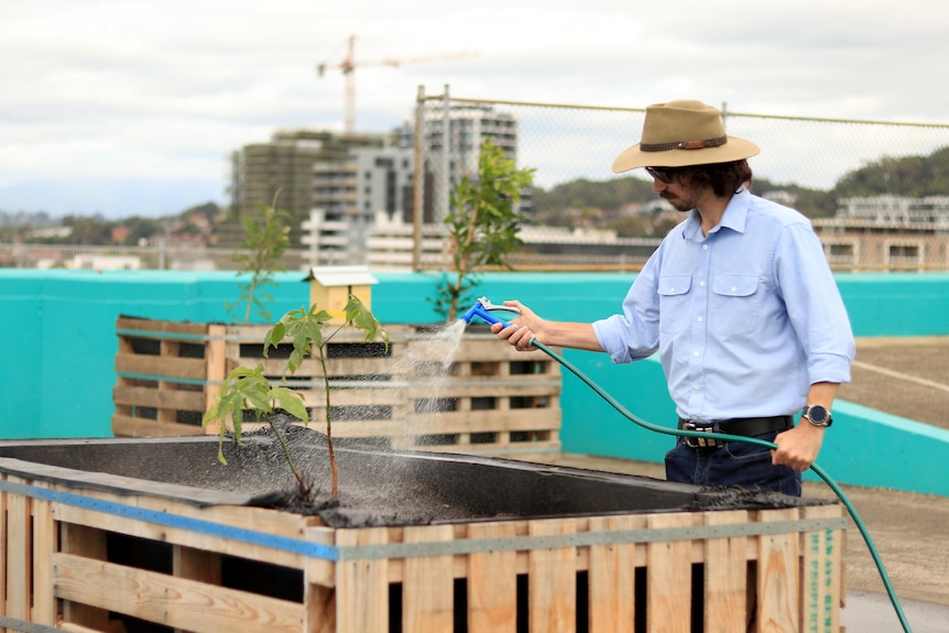 Jacob Williams wears a wide brimmed hat and blue shirt while watering plants in a car park.