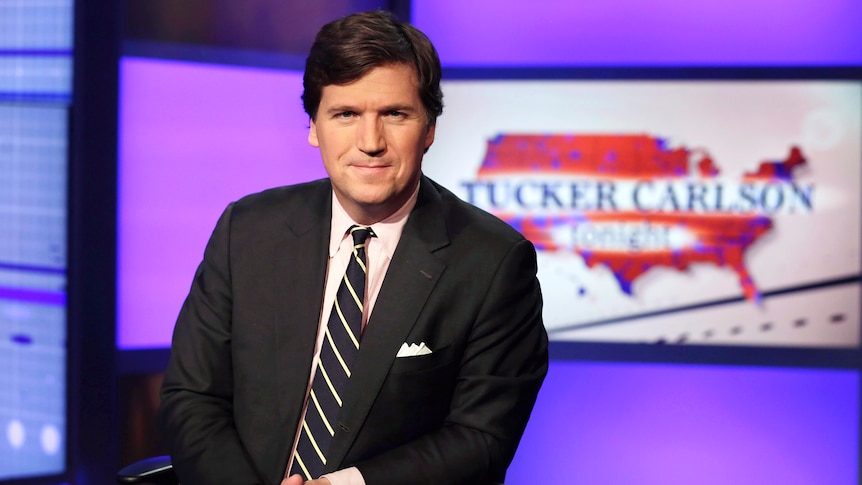 A man with brown hair is wearing a suit and tie in a tv-studio. Behind him a big screen saying  'Tucker Carlson tonight'.