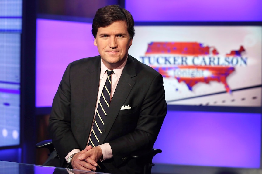 A man with brown hair is wearing a suit and tie in a tv-studio. Behind him a big screen saying  'Tucker Carlson tonight'.