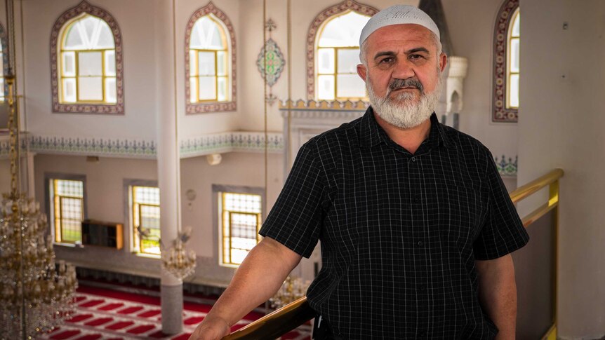 A man with a white camp stands on a balcony inside a mosque with decorated windows and bright red and white carpet.