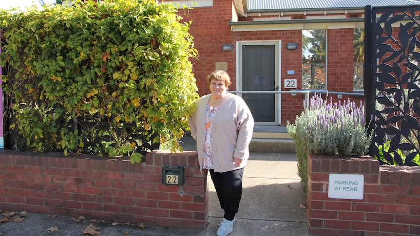 Woman in a cream cardigan standing next to a great hedge in front of a brick house