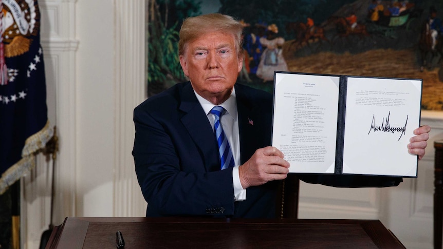 Donald Trump sits at a desk and holds up a signed presidential memorandum.