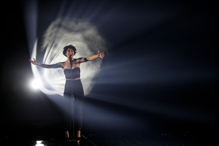 Seorang wanita berdiri di atas panggung yang gelap dengan cahaya terang di belakangnya.  Dia mengangkat lengannya dan bernyanyi di mikrofon, mata tertutup