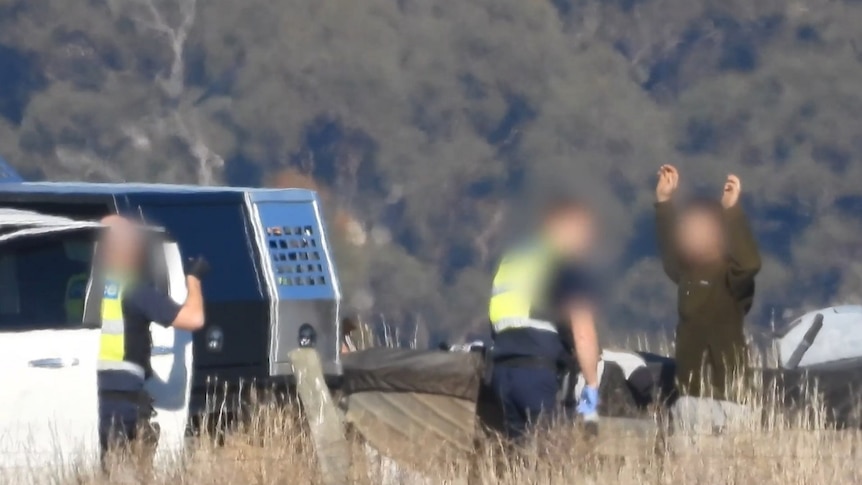 A man holds his hands up as to police officers with gloves inspect his boat.