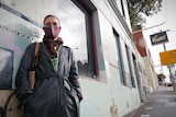 A young woman with a long black coat  and checked mask stands outside a pub