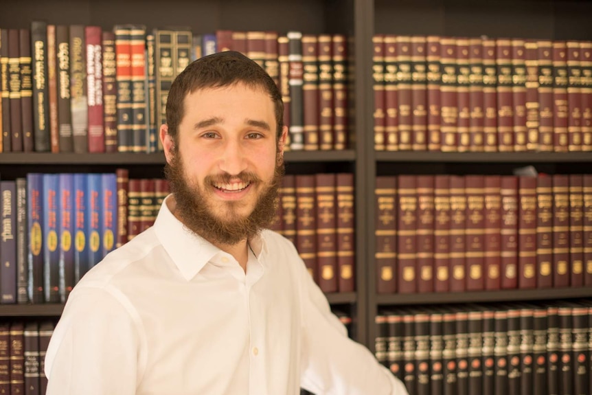 a man with a beard, stands, smiling in front of a bookshelf filled with Hebrew texts.