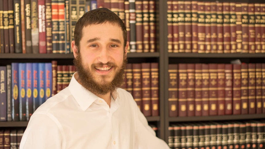 Rabbi Ari Rubin stands, smiling in front of a bookshelf filled with Hebrew texts.