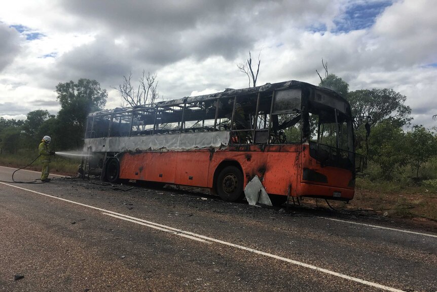 A burnt-out orange school bus lies on the side of the road with a firefighter hosing it down.