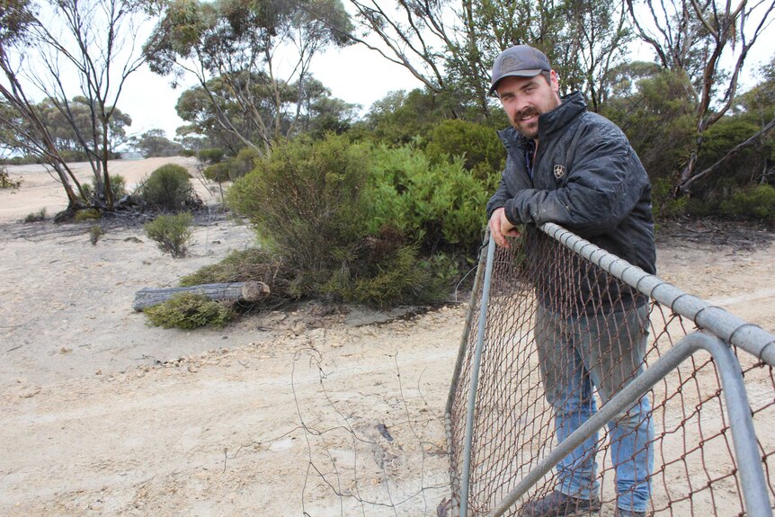 A man poses at a broken fence gate on an arid property.