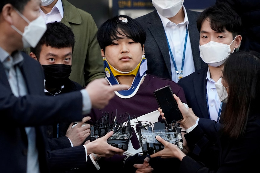 A boy with cropped black hair wearing a neck brace stands with people wearing masks.