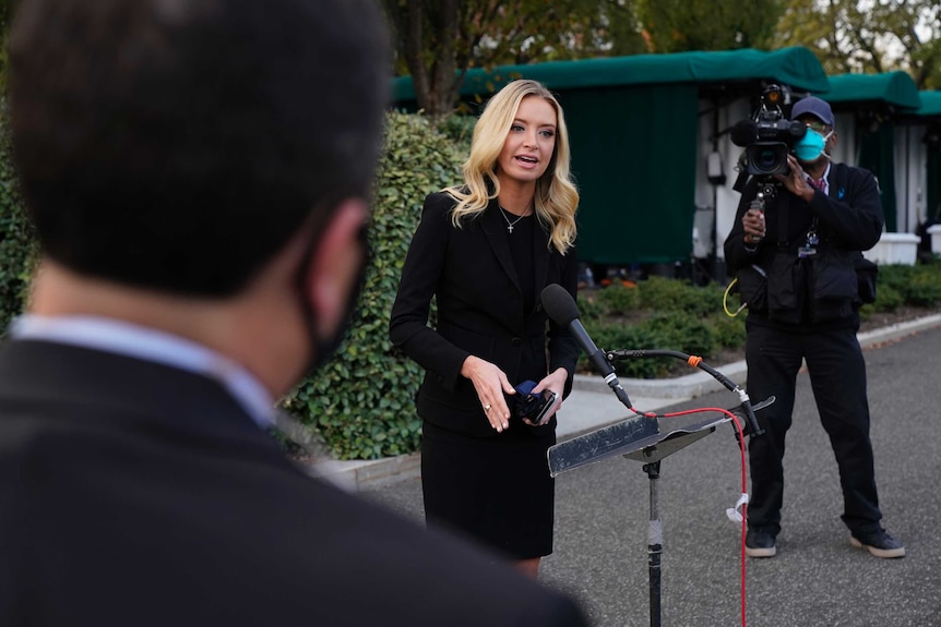 Kayleigh McEnany stands at a microphone wearing a black dress and no mask two men nearby are wearing masks.