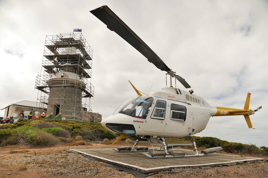 A helicopter sits on a helipad in front of a lighthouse on an island.