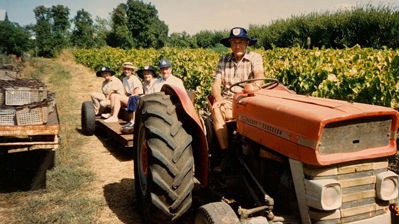 A man is wearing a hat, sitting on a tractor. It has a trailer behind it with four people in it.