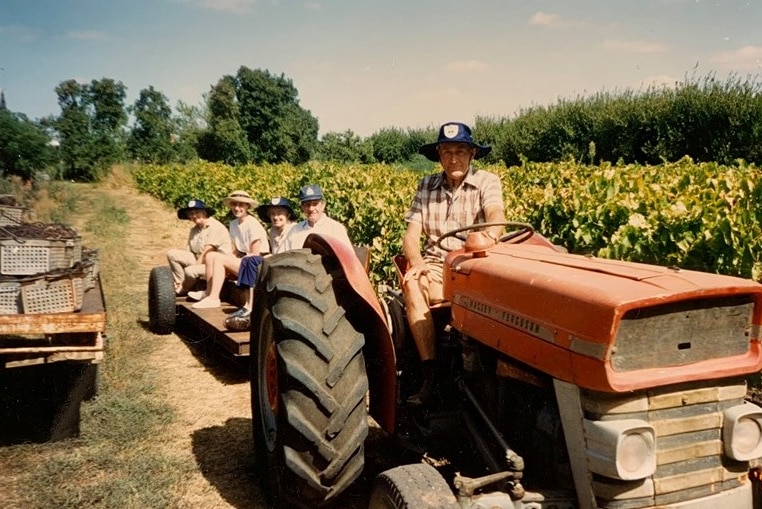 A man is wearing a hat, sitting on a tractor. It has a trailer behind it with four people in it.