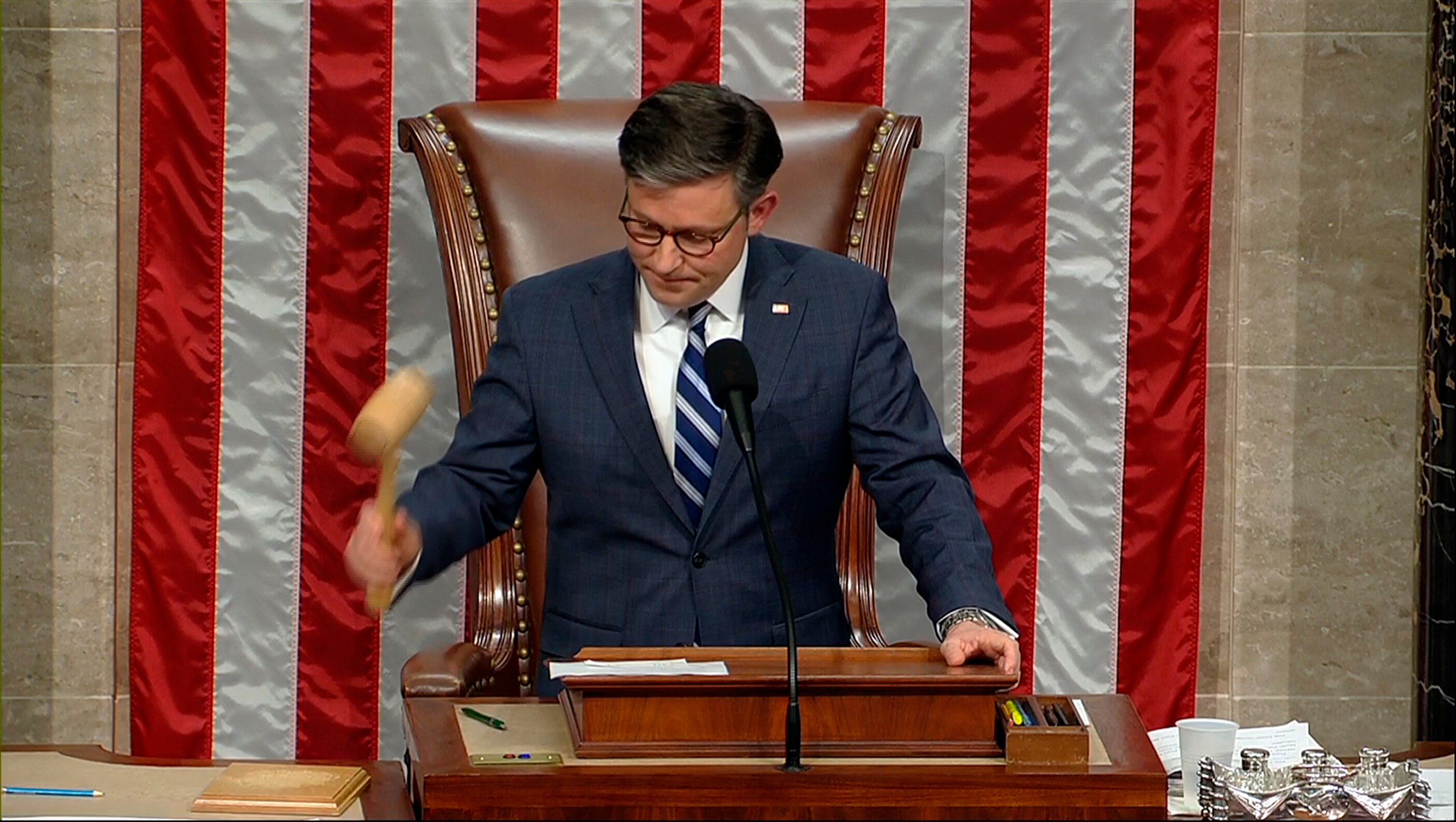 A man sitting in a chair lifts a gavel to bang it on his desk.