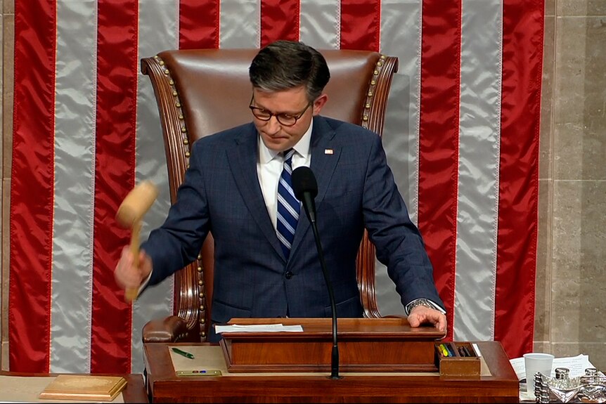 A man sitting in a chair lifts a gavel to bang it on his desk.