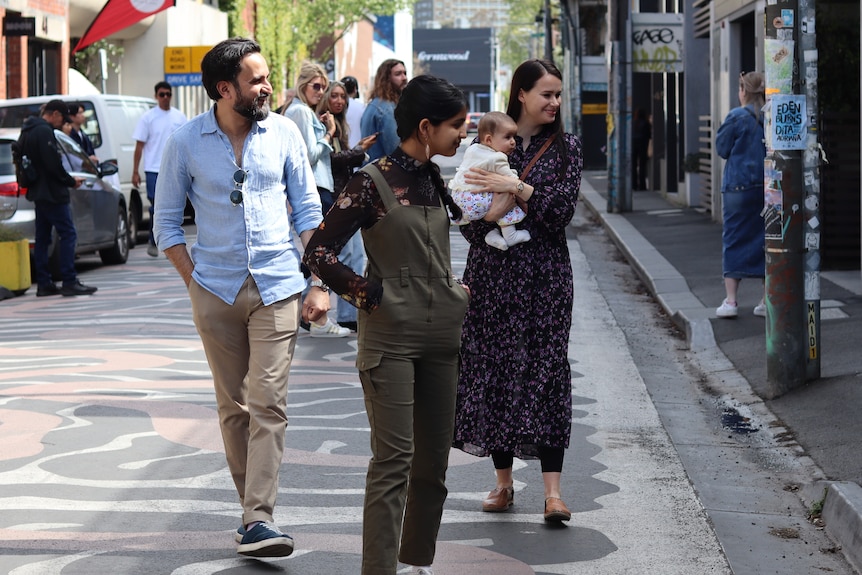 A family of a man and a woman holding a baby, and a teenaged girl, walk down a street.