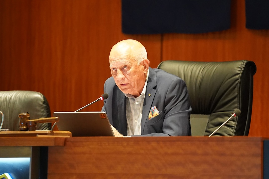 An older man in a suit sits in council chambers.