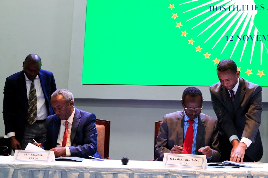 two people in chairs signing papers next to two people standing