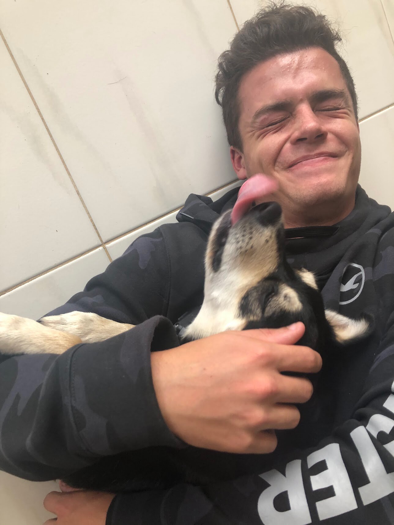 Ένας νεαρός άνδρας με κουκούλα χαμογελώντας και τσακίζει καθώς ένας σκύλος που κρατάει προσπαθεί να του γλείψει το πρόσωπό του.